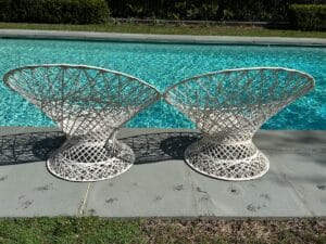 Two white, woven chairs on a stone patio beside a clear blue swimming pool with grass in the background.
