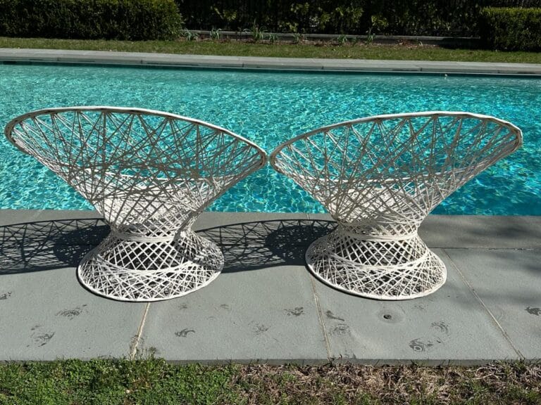 Two white, woven chairs on a stone patio beside a clear blue swimming pool with grass in the background.