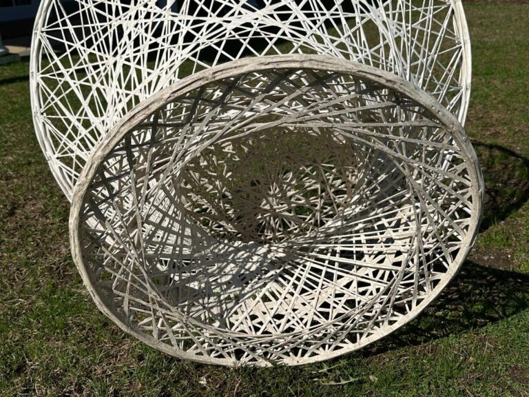 Woven circular sculpture with intricate patterns, placed on grass, casting shadows.