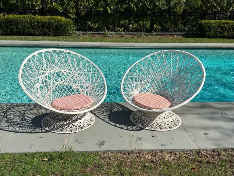 Two white, woven lounge chairs with red cushions beside a sparkling blue pool, set on a sunny patio.