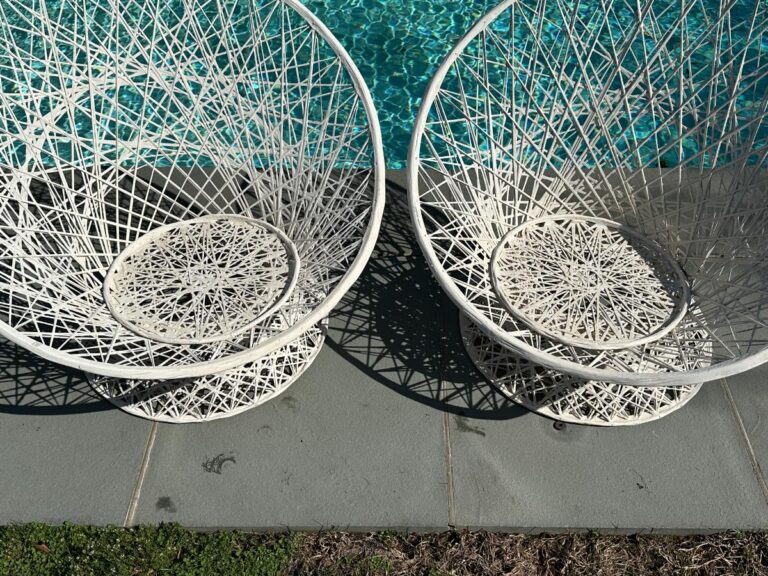 Two white, round wicker chairs by a pool on a sunny day, casting shadows on the gray pavement.