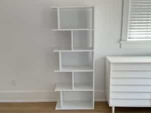 A modern white bookshelf with staggered shelves stands next to a white dresser in a bright room.