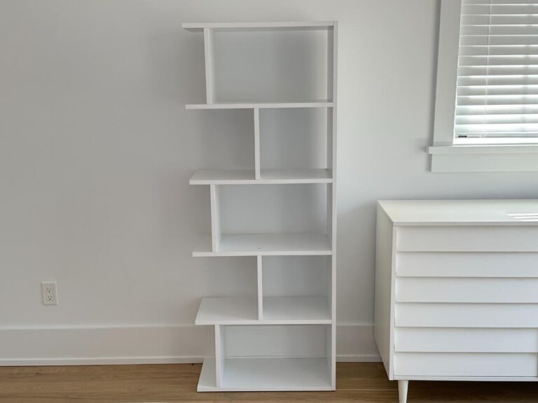 A modern white bookshelf with staggered shelves stands next to a white dresser in a bright room.