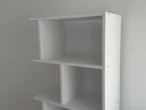 White, partially assembled bookshelf with three shelves against a plain, light-colored wall.