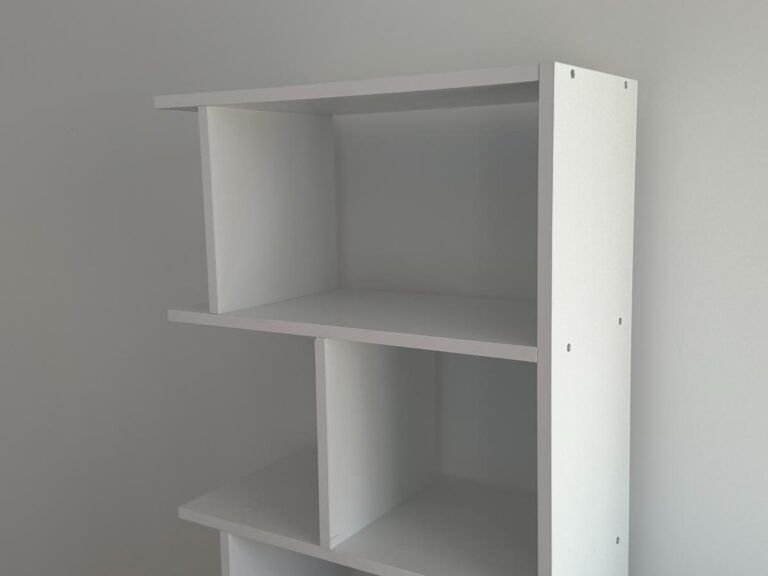 White, partially assembled bookshelf with three shelves against a plain, light-colored wall.