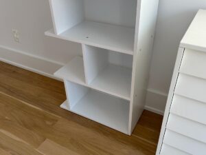 A white, partially assembled shelf unit on a wooden floor next to a white chest of drawers.