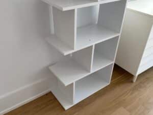 White modern shelving unit with open compartments, standing on a wooden floor next to a white cabinet.