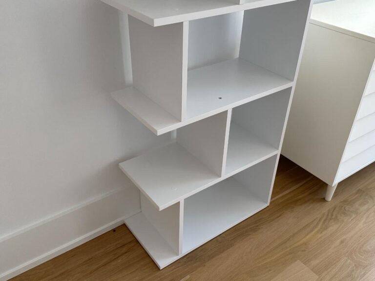 White modern shelving unit with open compartments, standing on a wooden floor next to a white cabinet.