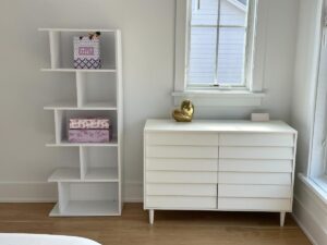 White bookshelf with decorative boxes beside a white dresser and window in a bright, modern room.