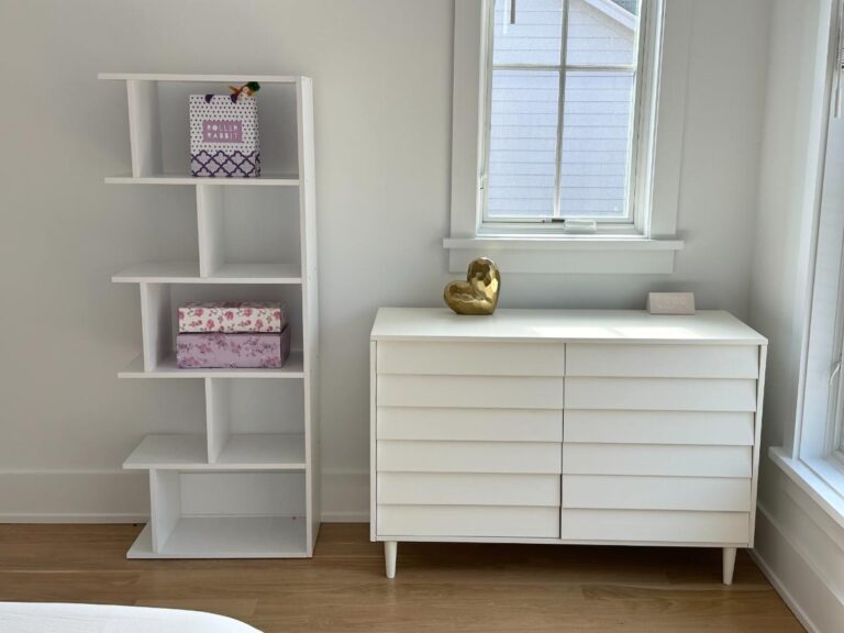 White bookshelf with decorative boxes beside a white dresser and window in a bright, modern room.