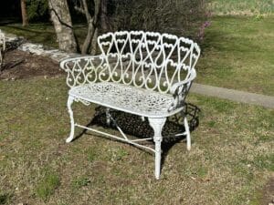 Ornate white metal bench with heart patterns, placed on grass in a sunlit garden setting.
