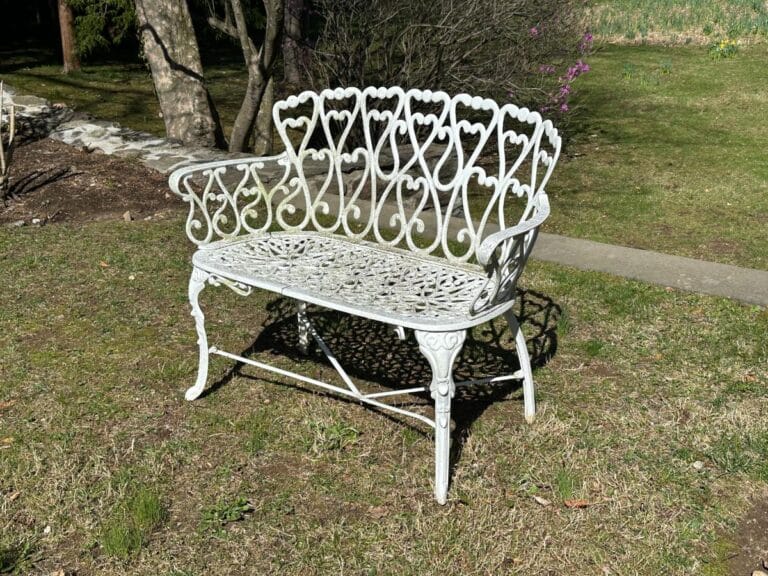 Ornate white metal bench with heart patterns, placed on grass in a sunlit garden setting.
