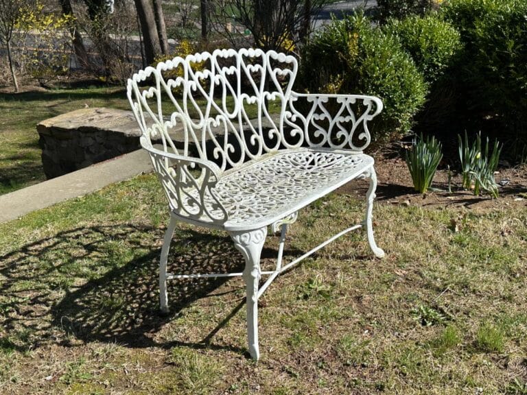 White ornate metal bench on grass, casting intricate shadows, with bushes and stone wall in the background.