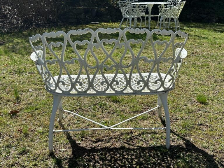 Ornate white metal bench with a heart-patterned backrest on a grassy lawn, with white chairs and table in the background.