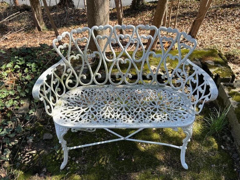 Ornate white metal bench with intricate heart pattern in a forest setting.