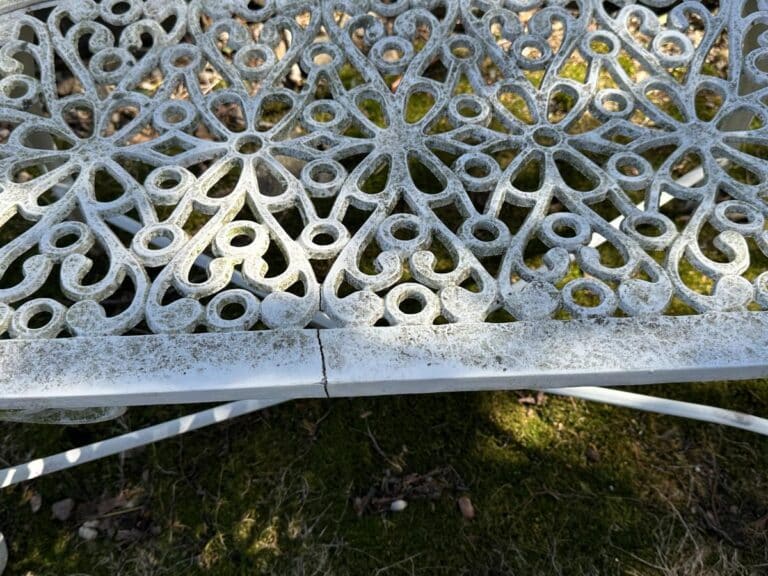 Close-up of an ornate, weathered metal chair with a crack, set on a grassy background.