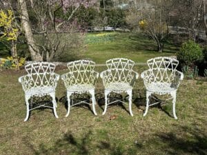 Four ornate white metal chairs on a grassy lawn, surrounded by trees and spring blooms.