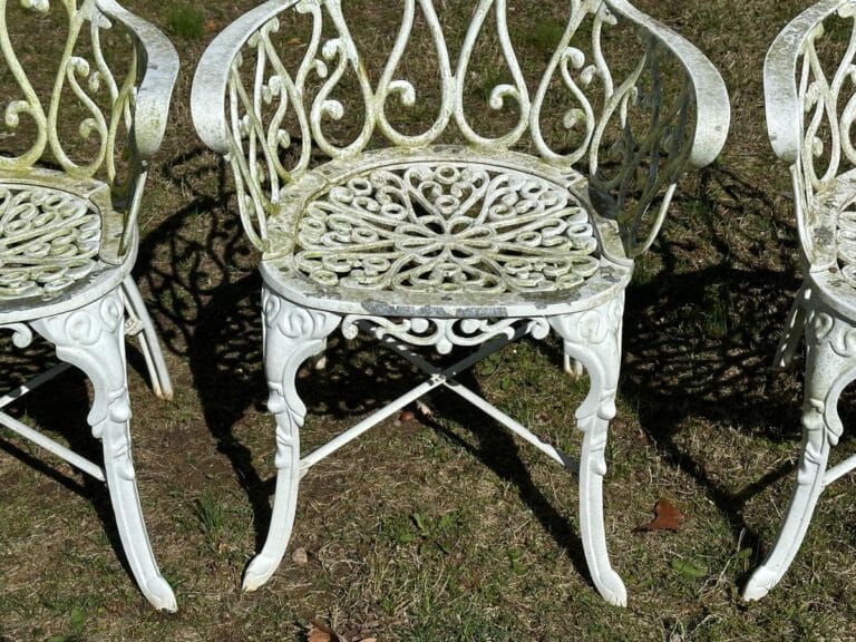 Three ornate white wrought-iron chairs with intricate patterns on grass, showing some weathering and moss.