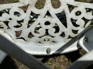 Close-up of ornate white metalwork on a garden chair with light rust spots and grass visible below.
