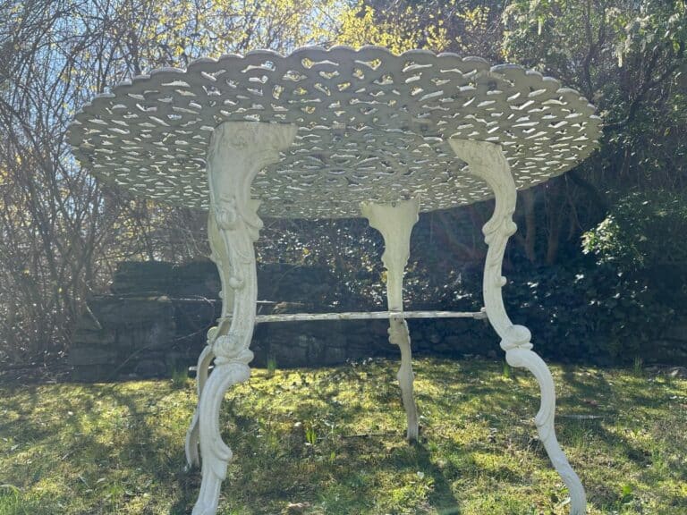 Ornate, white metal table with intricate design on grass in a sunlit garden setting.