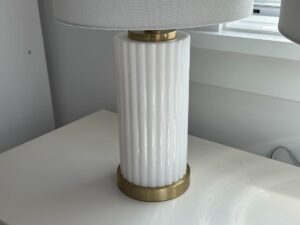 White ribbed ceramic table lamp with gold accents on a white surface near a window.
