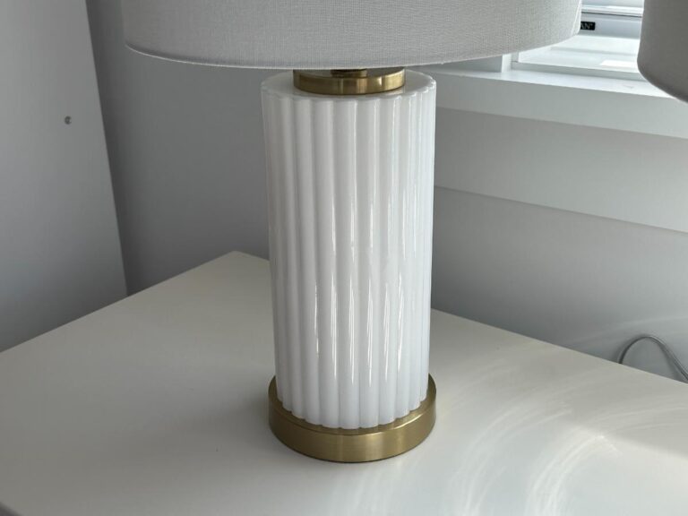 White ribbed ceramic table lamp with gold accents on a white surface near a window.