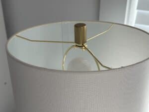 White lampshade with brass hardware and a visible light bulb inside, near a white window frame.