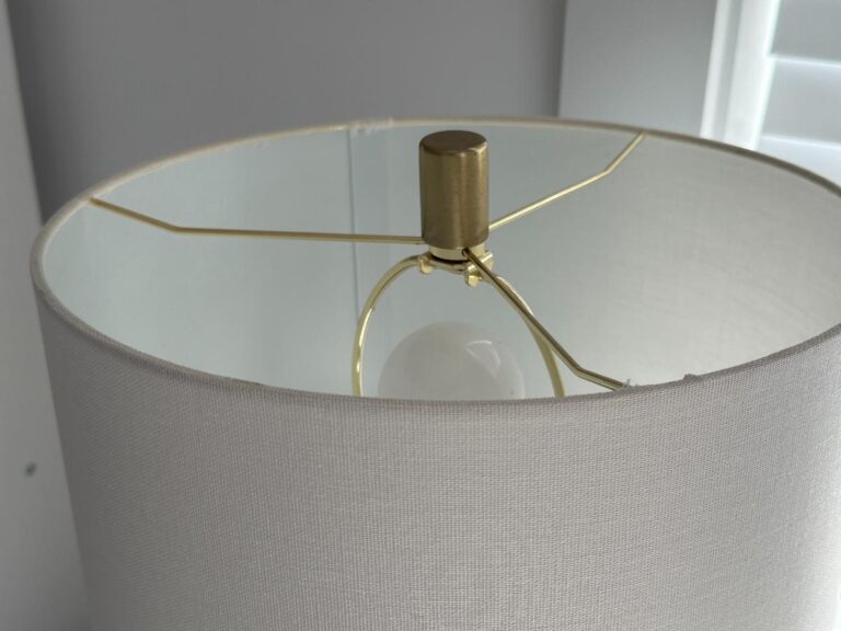 White lampshade with brass hardware and a visible light bulb inside, near a white window frame.