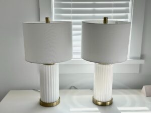Two white table lamps with gold accents on a white desk in front of a window with white blinds.