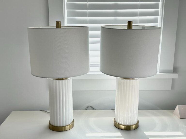 Two white table lamps with gold accents on a white desk in front of a window with white blinds.